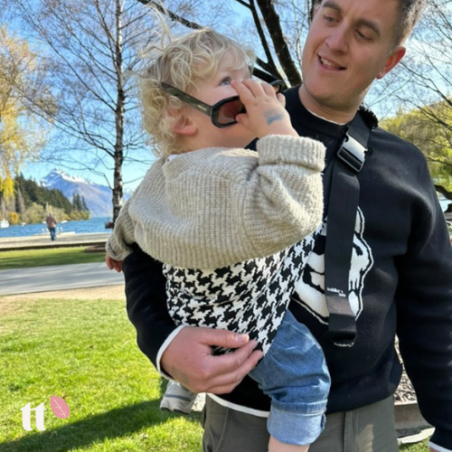 A toddler is being carried in a black and white houndstooth toddler tote, which is worn on the chest. The carrier has an adjustable strap with a shoulder pad, and it appears to be made of a soft fabric.