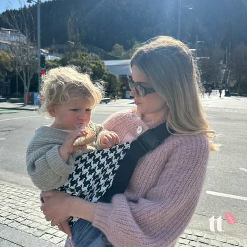 A toddler is being carried in a black and white houndstooth toddler tote, which is worn on the chest. The carrier has an adjustable strap with a shoulder pad, and it appears to be made of a soft fabric.