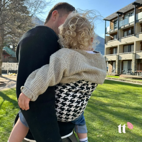 A toddler is being carried in a black and white houndstooth toddler tote, which is worn on the chest. The carrier has an adjustable strap with a shoulder pad, and it appears to be made of a soft fabric.